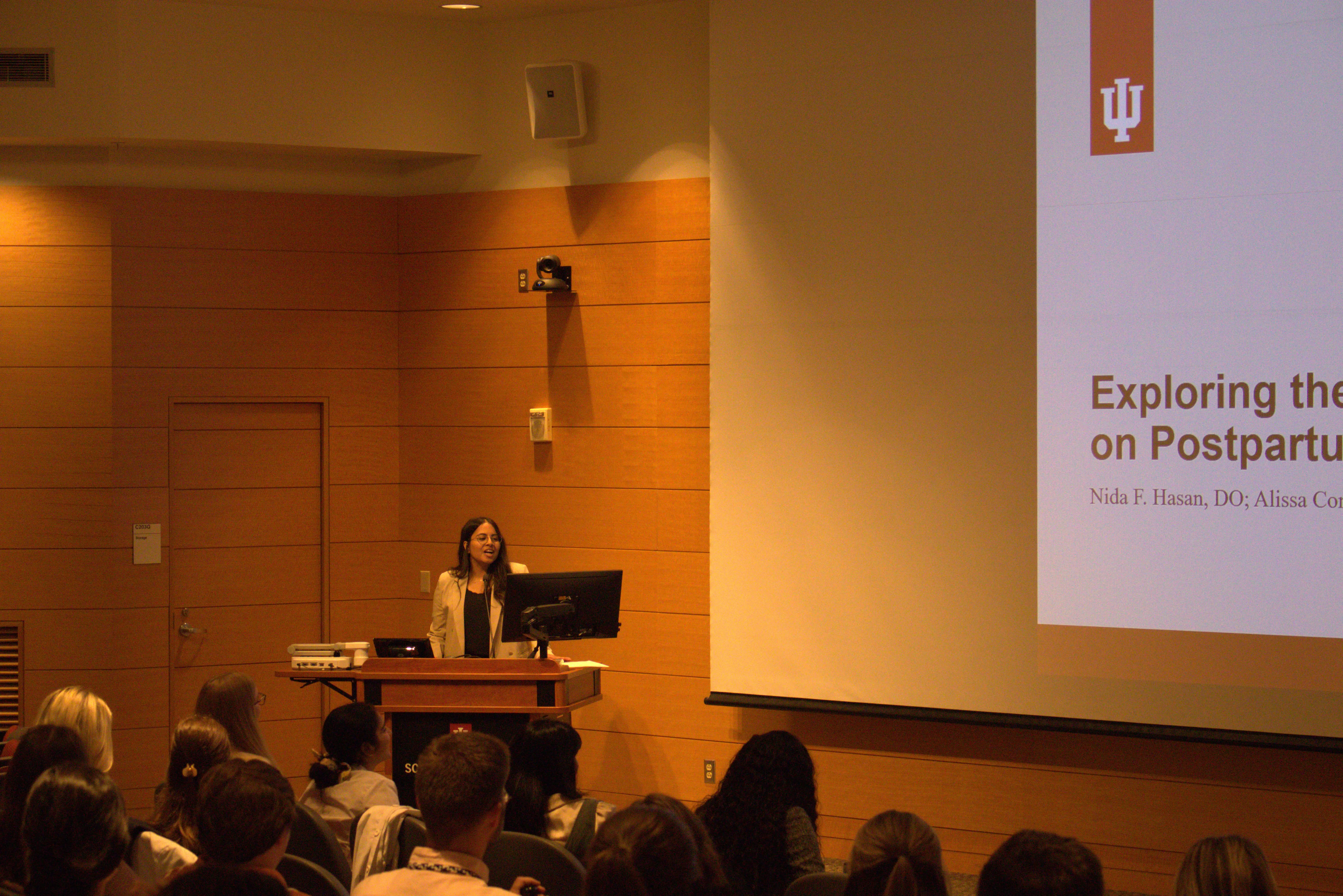 Dr. Nida Hasan stands at the lectern in front of her audience in an auditorium
