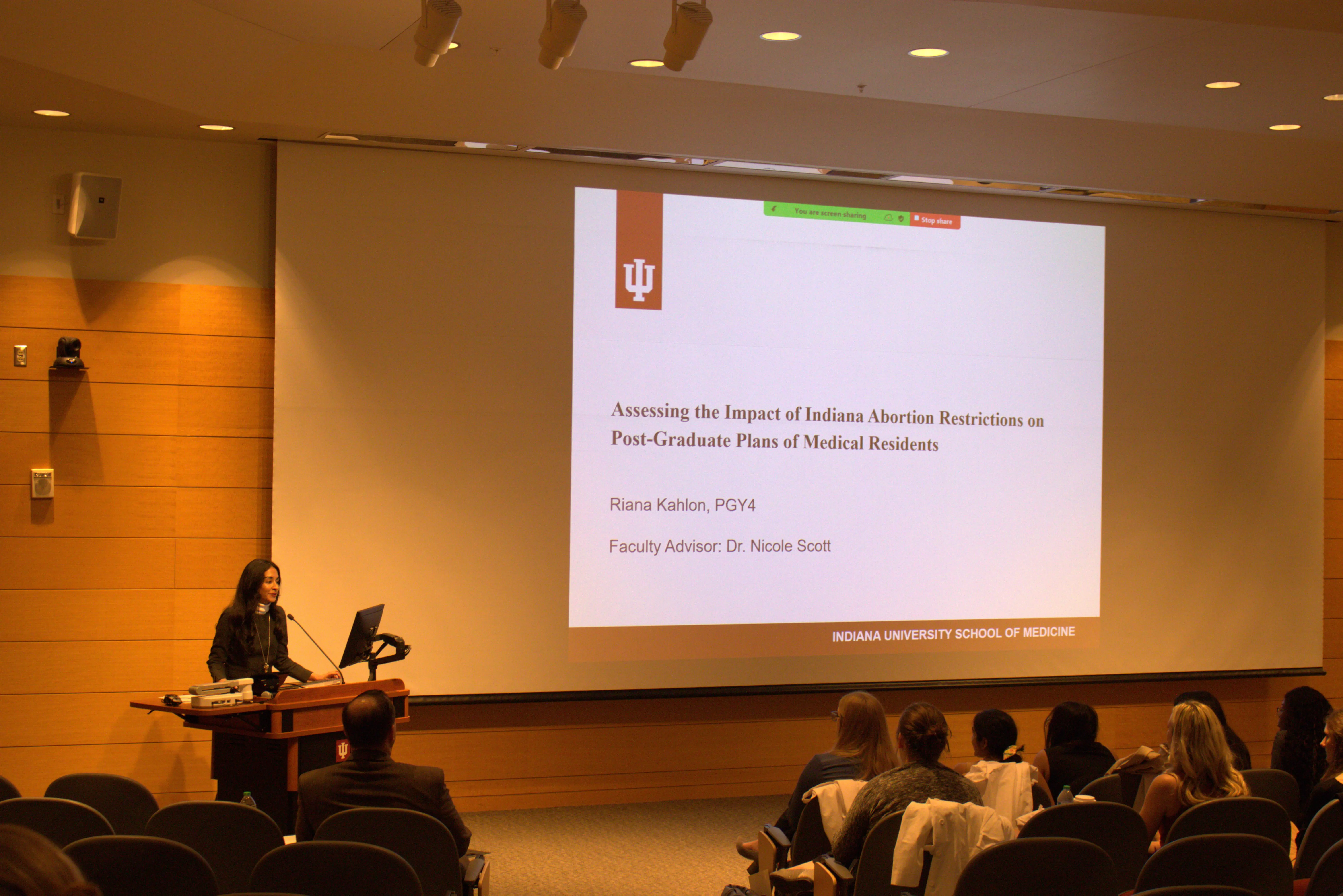 Dr. Riana Kahlon stands at the lectern facing the audience in the auditorium