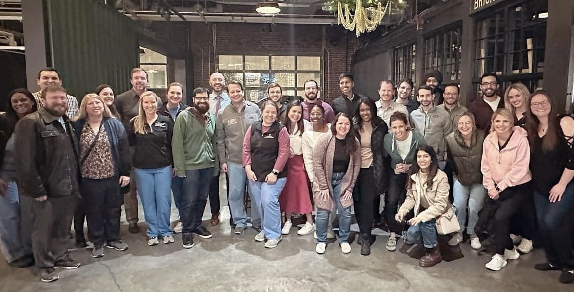 a large group of about 30 trainees and faculty stand together and smile at a celebratory gathering