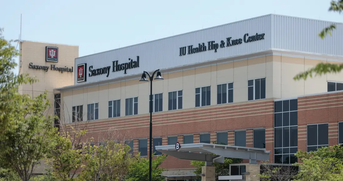 A photo shows the exterior of Saxony Hospital on a sunny day. Trees frame the sides of the photo. Text on the building reads "Saxony Hospital. IU Health Hip & Knee Center." 