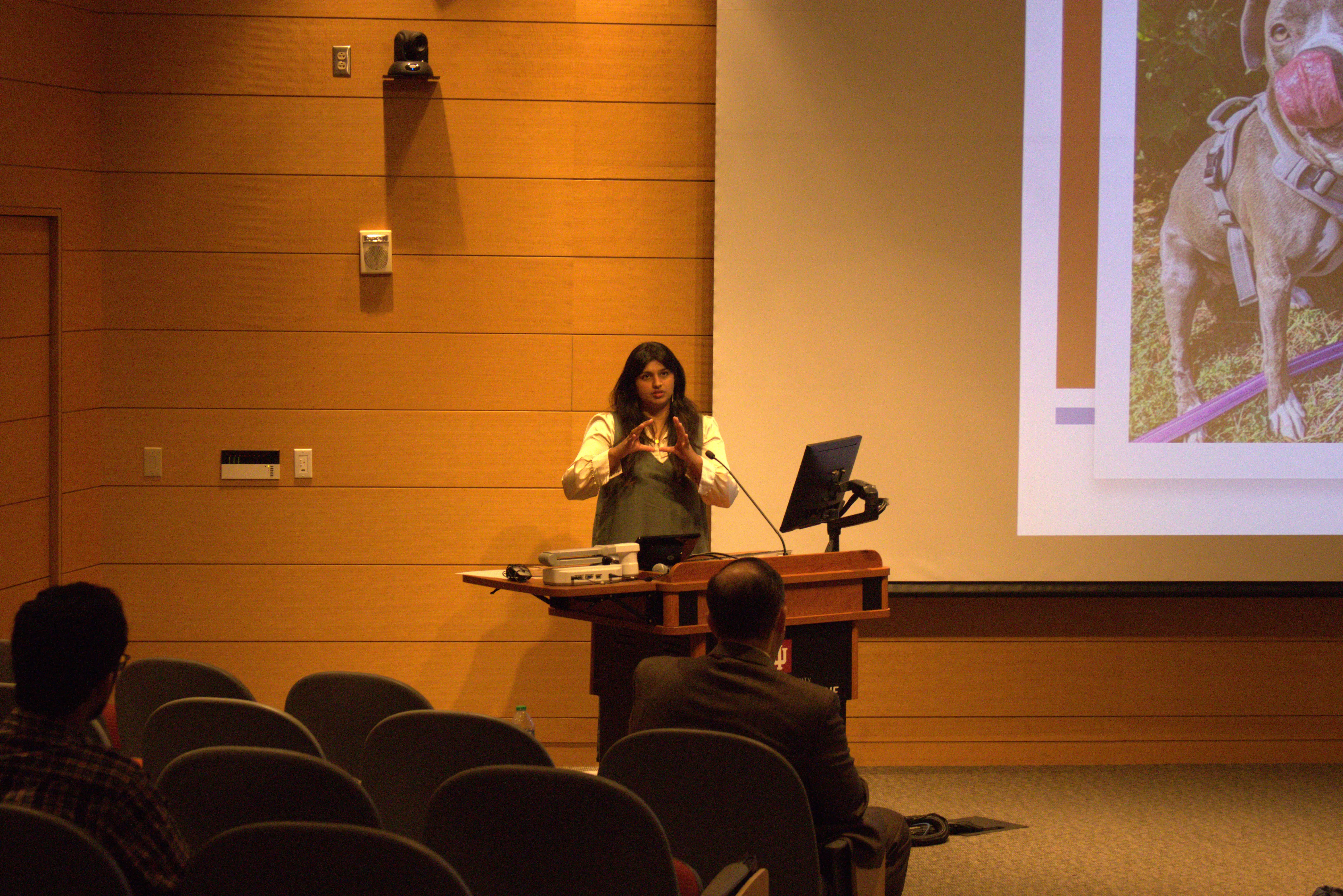 Dr. Prakrithi Srinand stands at the lectern gesturing while the audience watches