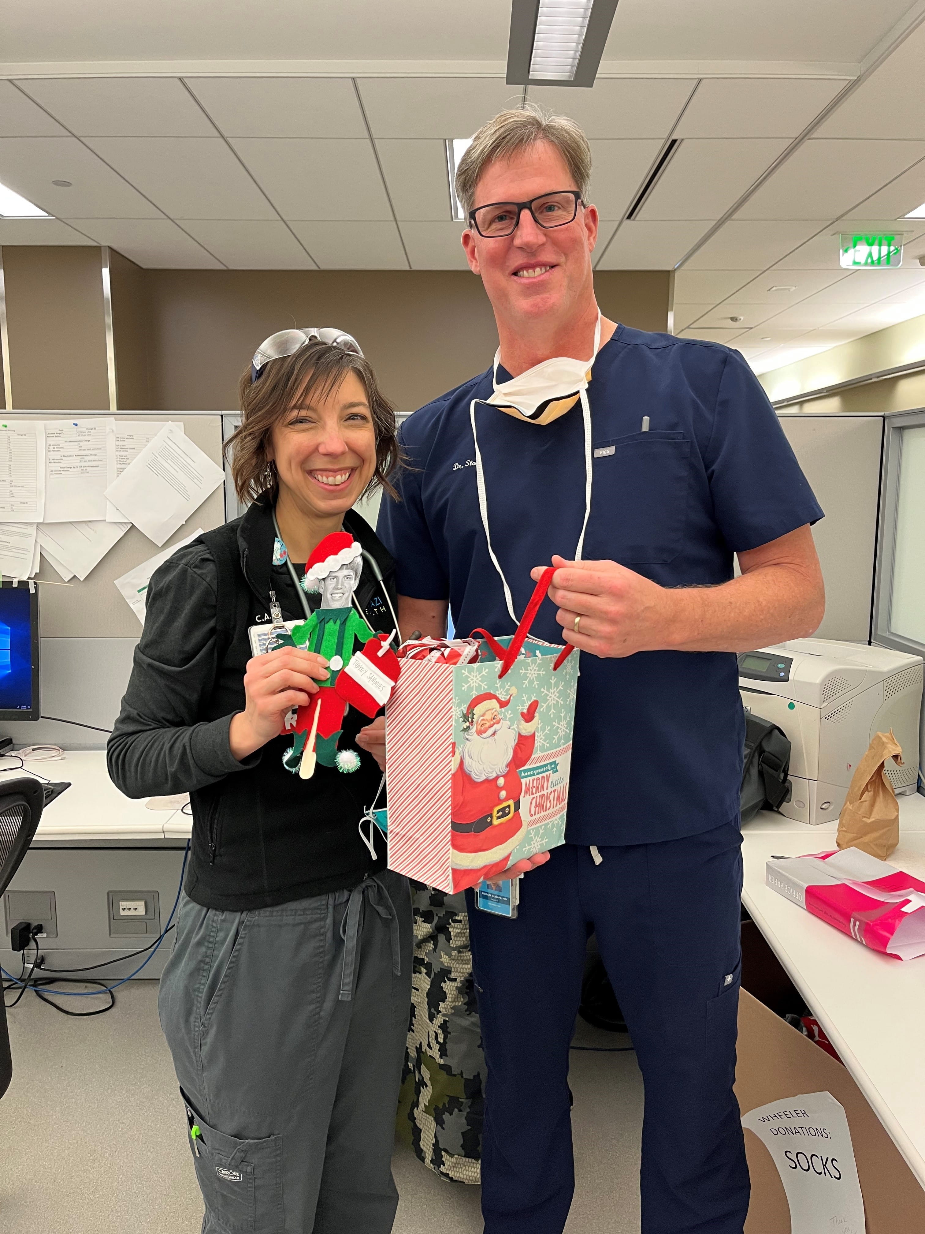 brian sloan and a colleague hold a gift bag with santa on it in the breakroom. They are both wearing scrubs.