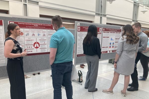 A group of students and observers having a discussion in front of a set of poster boards showing research projects.