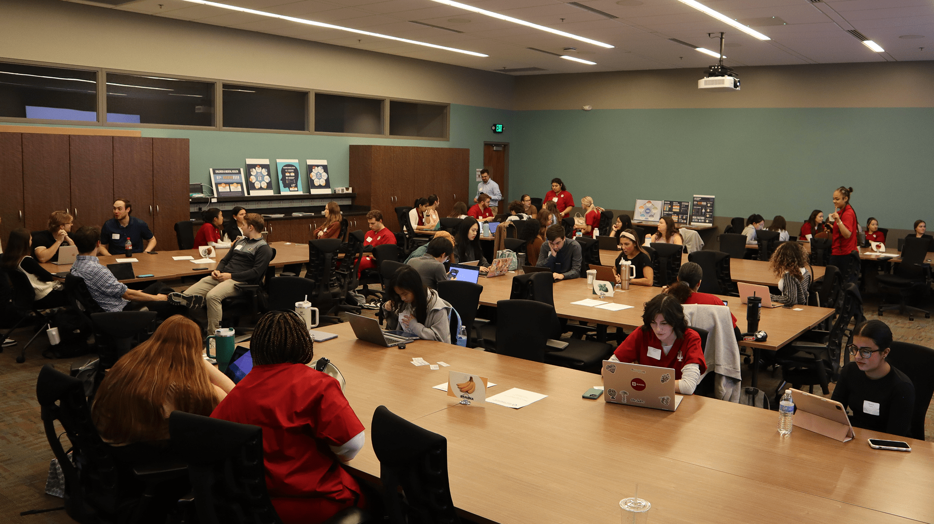Large room with people seated at tables engaged in discussions or working on laptops, with posters and a ceiling-mounted projector visible.