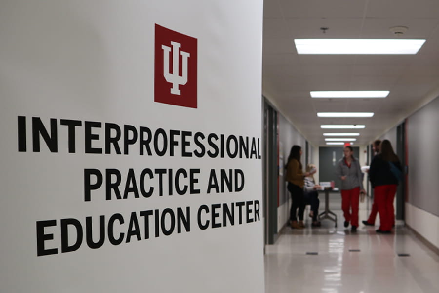A large white banner in the foreground prominently displays the red Indiana University (IU) trident logo and the black text 'INTERPROFESSIONAL PRACTICE AND EDUCATION CENTER'. In the background, a brightly lit modern hallway stretches into the distance, with several blurred figures of people, likely students or staff, interacting or walking, suggesting activity within the center. The scene implies a vibrant educational or professional environment focused on interdisciplinary collaboration.
