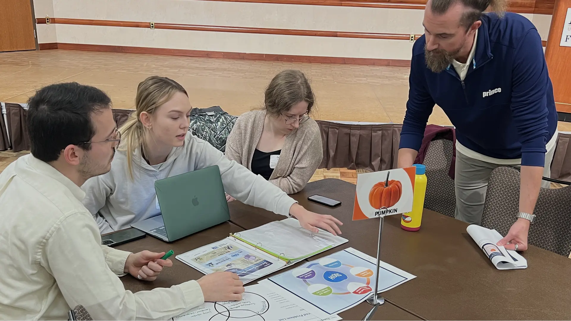 Four individuals seated around a table engaged in discussion with documents, diagrams, and a laptop, including a labeled “Pumpkin” diagram.