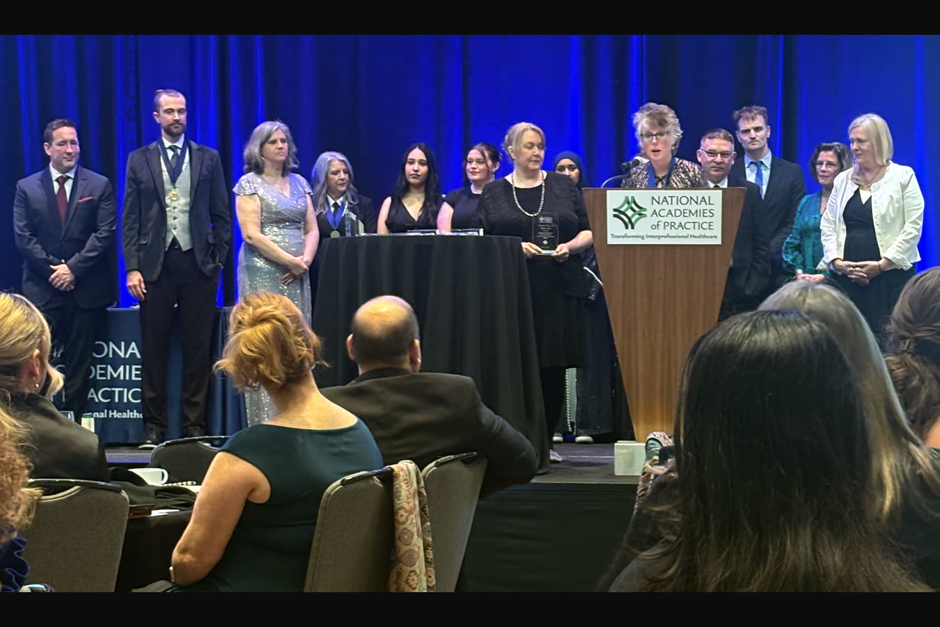 April stands at a podium during the National Academies of Practice awards ceremony while members of the Comprehensive Pain Assessment Clinic and Indiana University Interprofessional Practice and Education Center teams stand behind her on stage as the Creativity in Practice & Education Award is presented.