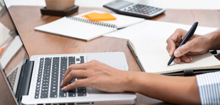 Person working on a laptop and writing notes in a notebook at a wooden desk with office supplies, calculator, and notepad – productivity and remote work concept.