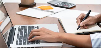 Person working on a laptop and writing notes in a notebook at a wooden desk with office supplies, calculator, and notepad – productivity and remote work concept.