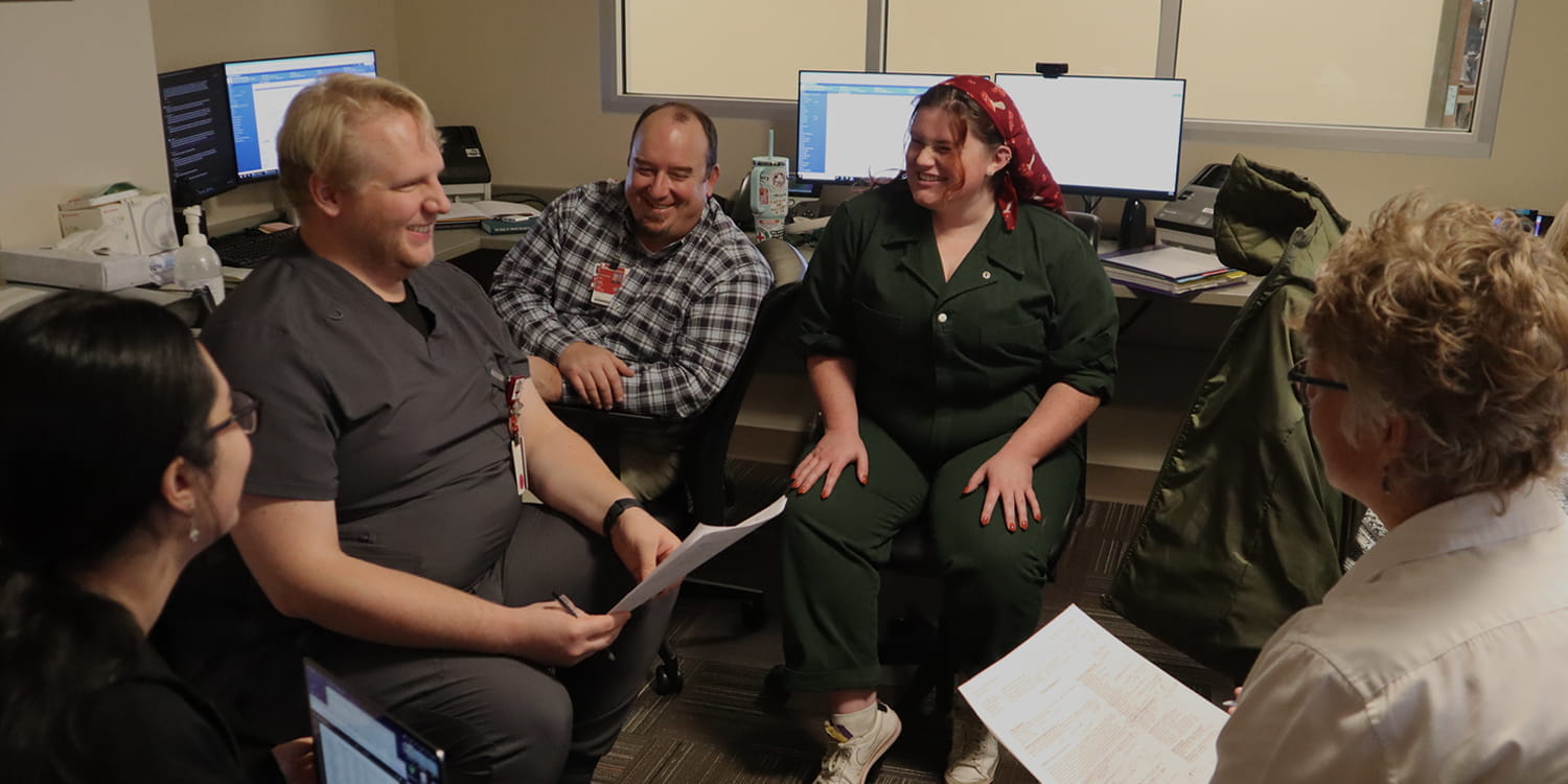 Group of six people in an office setting engaged in a meeting, with computer monitors and papers visible.