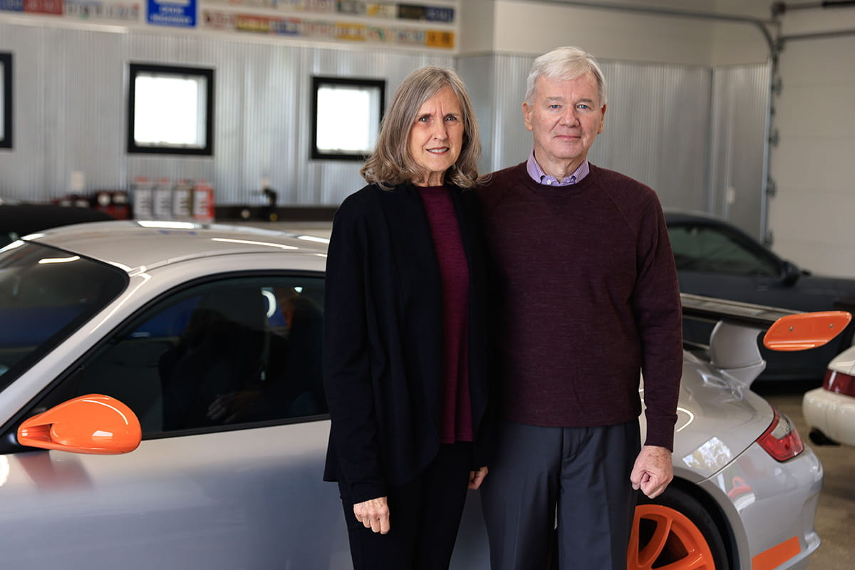 an older white couple stands in front of a luxury car