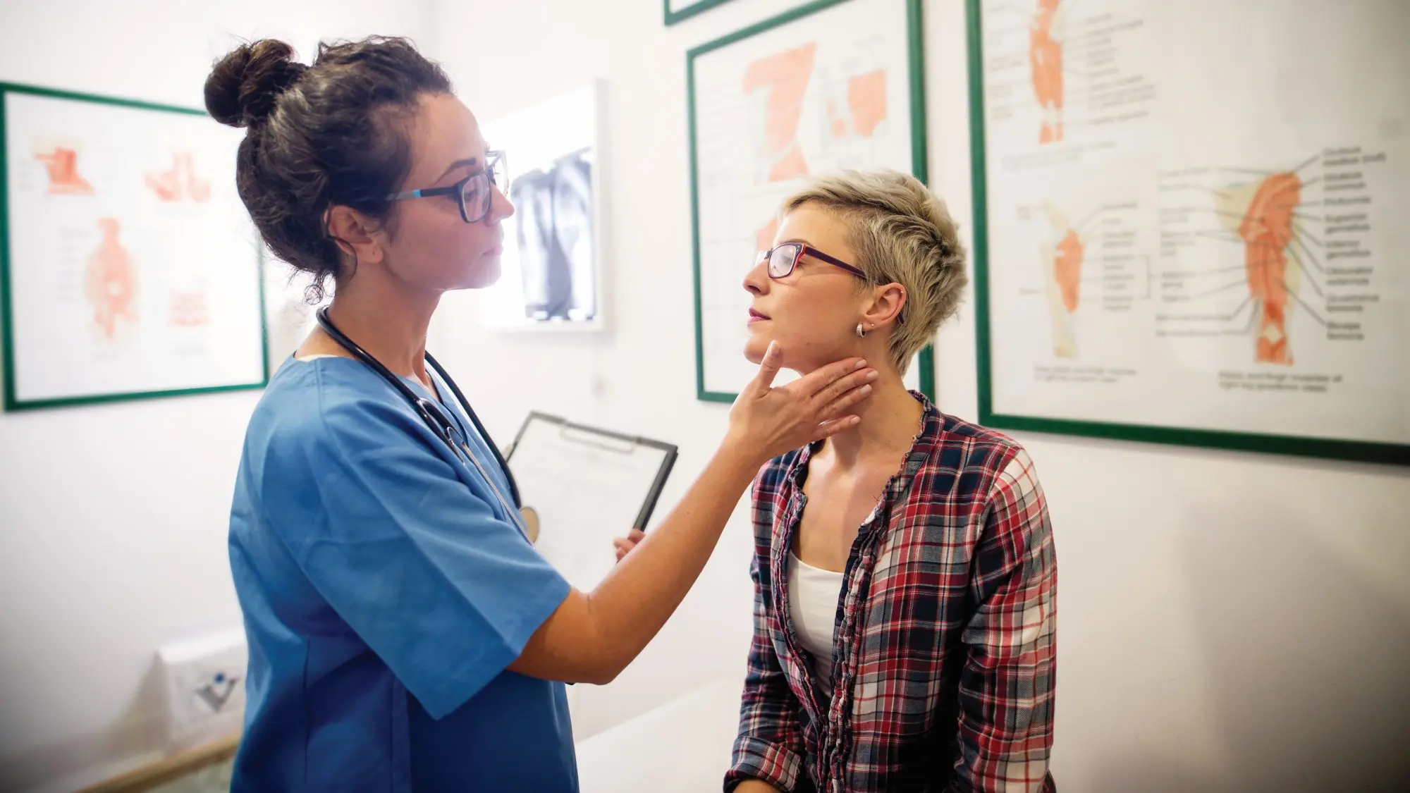 A health care provider wearing blue scrubs and a stethoscope with her hair in a loose high bun feels a patient's neck and lymph nodes in an exam room. 
