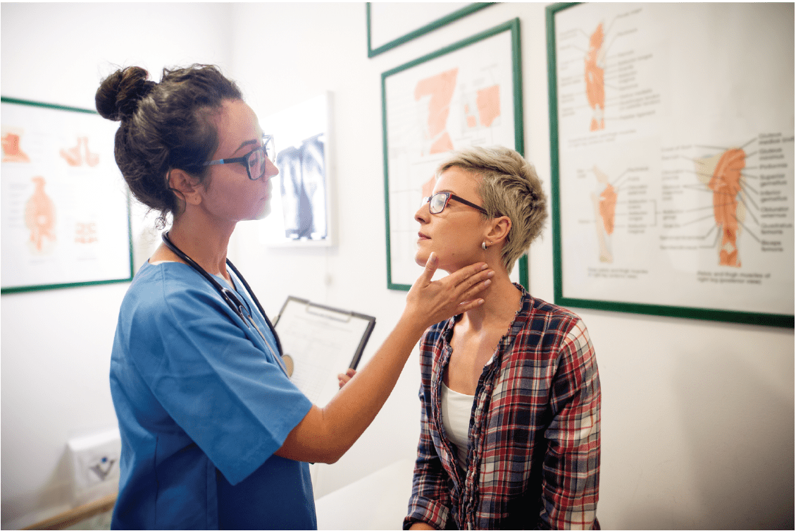 A health care provider wearing blue scrubs and a stethoscope with her hair in a loose high bun feels a patient's neck and lymph nodes in an exam room.