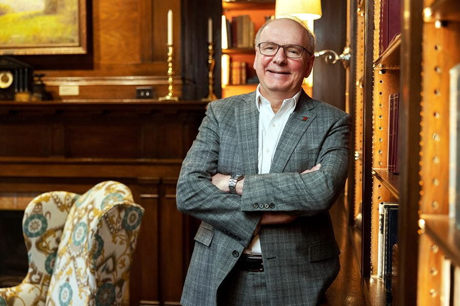 dean hess stands with his arms folder leaning against a shelf in his home library