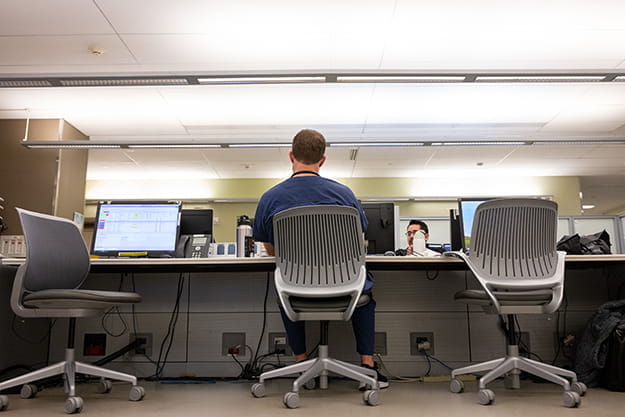 a physician in blue scrubs sits alone at a computer work station in the hospital. There are empty stations on either side of him.