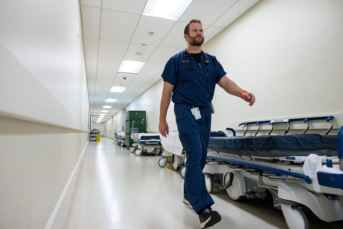Matt Rutz walks down a hospital hallway filled with hospital beds on wheels. He is wearing dark blue scrubs.