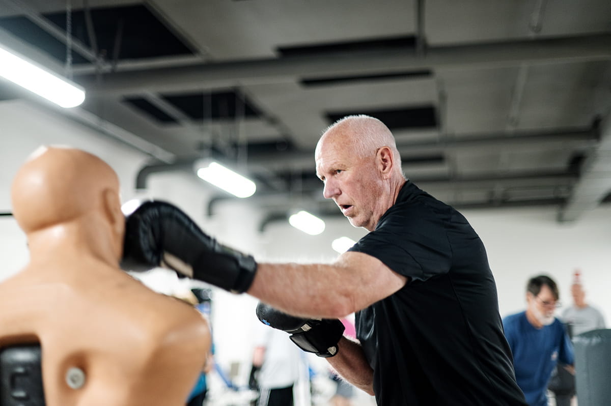 Ted Kitchel boxes against a dummy in the gym. He has short cropped white hair, a black t shirt and black boxing gloves.