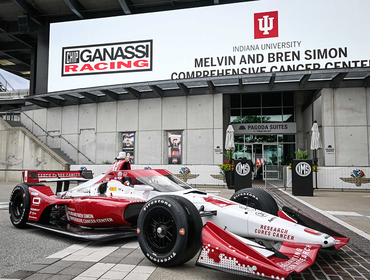 a red and white indy car race car is parked in front of the pagoda suites at the indianapolis motor speedway. A billboard in the background reads "Chip Ganassi Racing" and "Indiana University Melvin and Bren Simon Comprehensive Cancer Center"