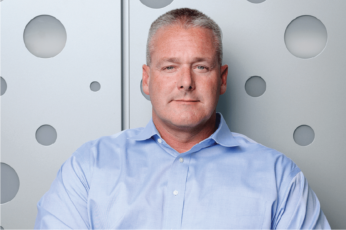 Kevin Smith sits in front of a metallic wall with circle detailing. He has short cropped gray hair, a blue button up shirt and a stoic expression.