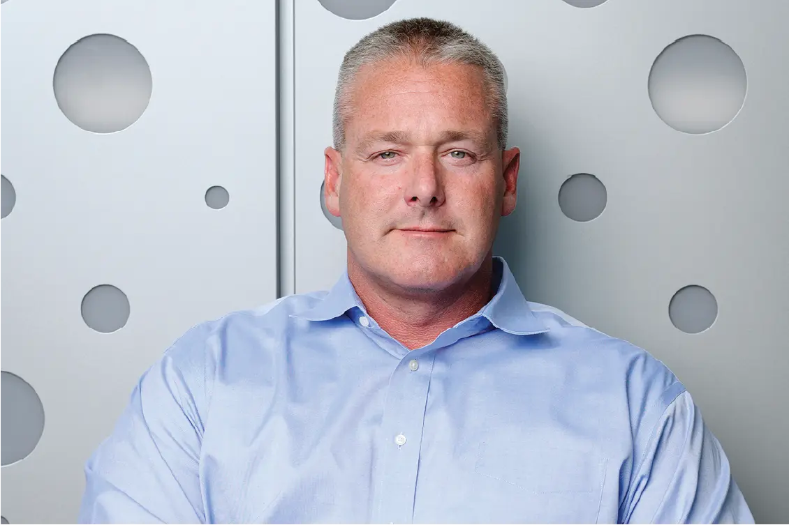 Kevin Smith sits in front of a metallic wall with circle detailing. He has short cropped gray hair, a blue button up shirt and a stoic expression.