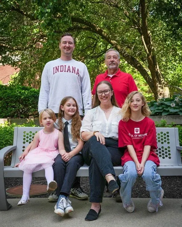 taylor etchison and her children, husband and father sit and stand around a bench outside on a sunny day