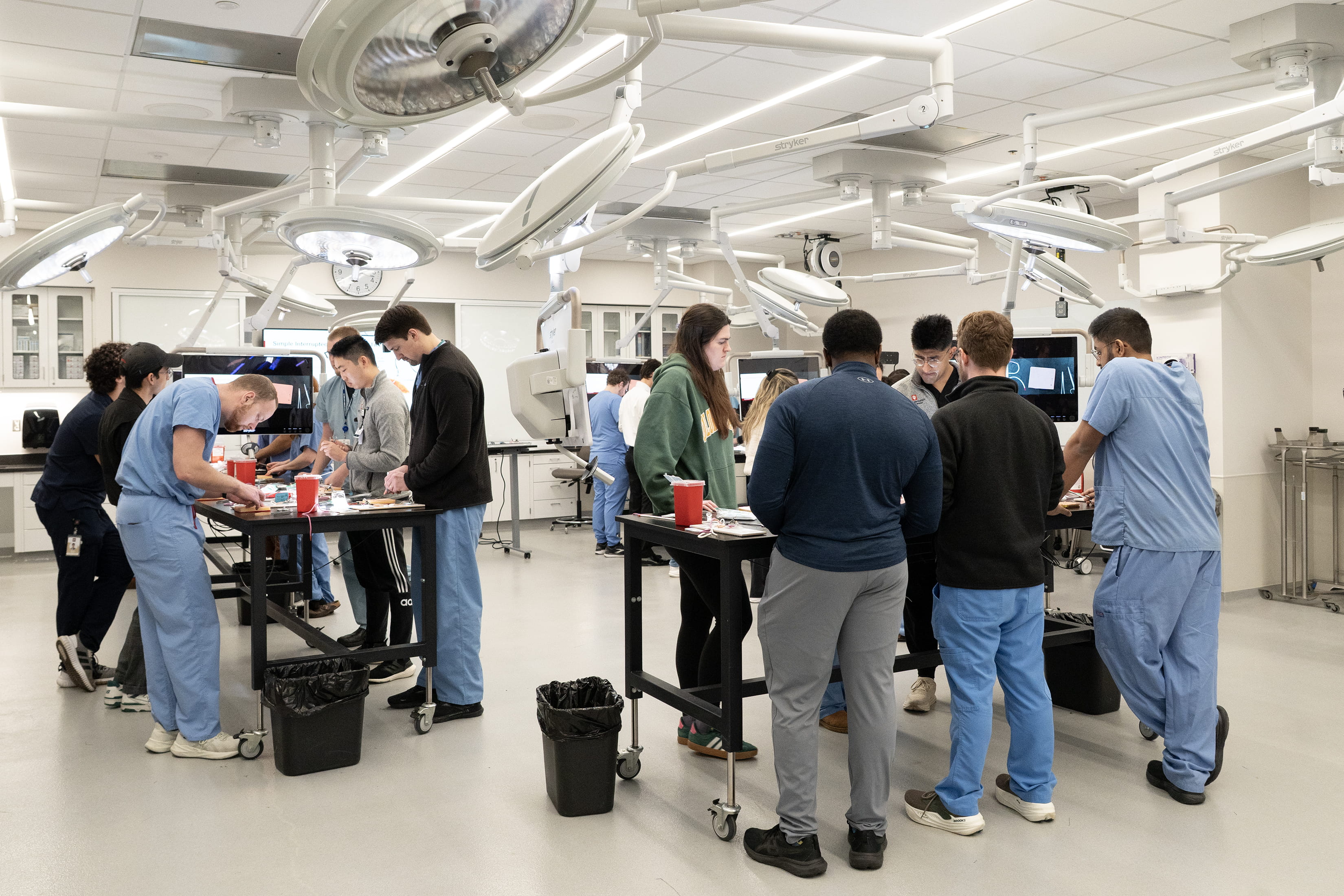 A dozen trainees work at standing tables in the surgical skills lab