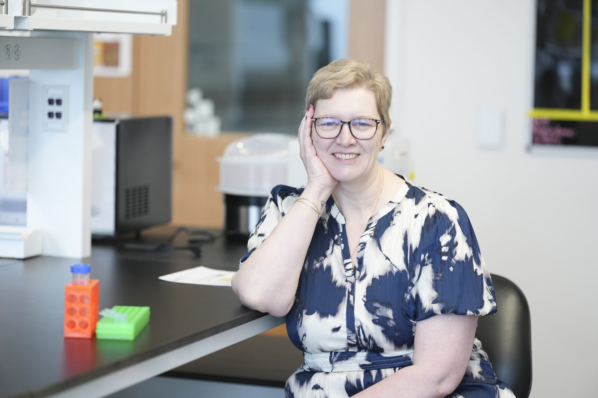 A scientist sitting on chair with her hand on her face smiling at the camera
