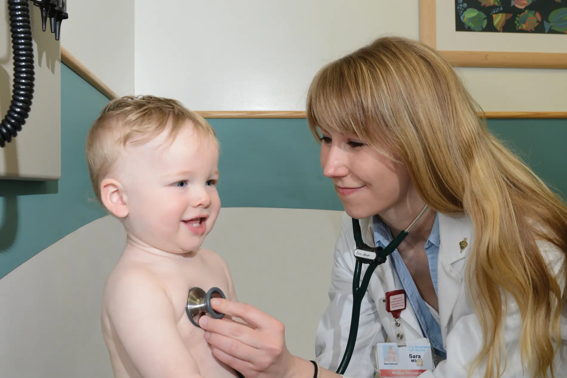 a pediatrician with long blond hair listens to the chest of a smiling toddler with her stethoscope