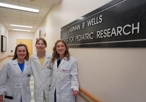 Three female MMIA students in white lab coats pose next a Herman B Wells Center for Pediatric Research sign