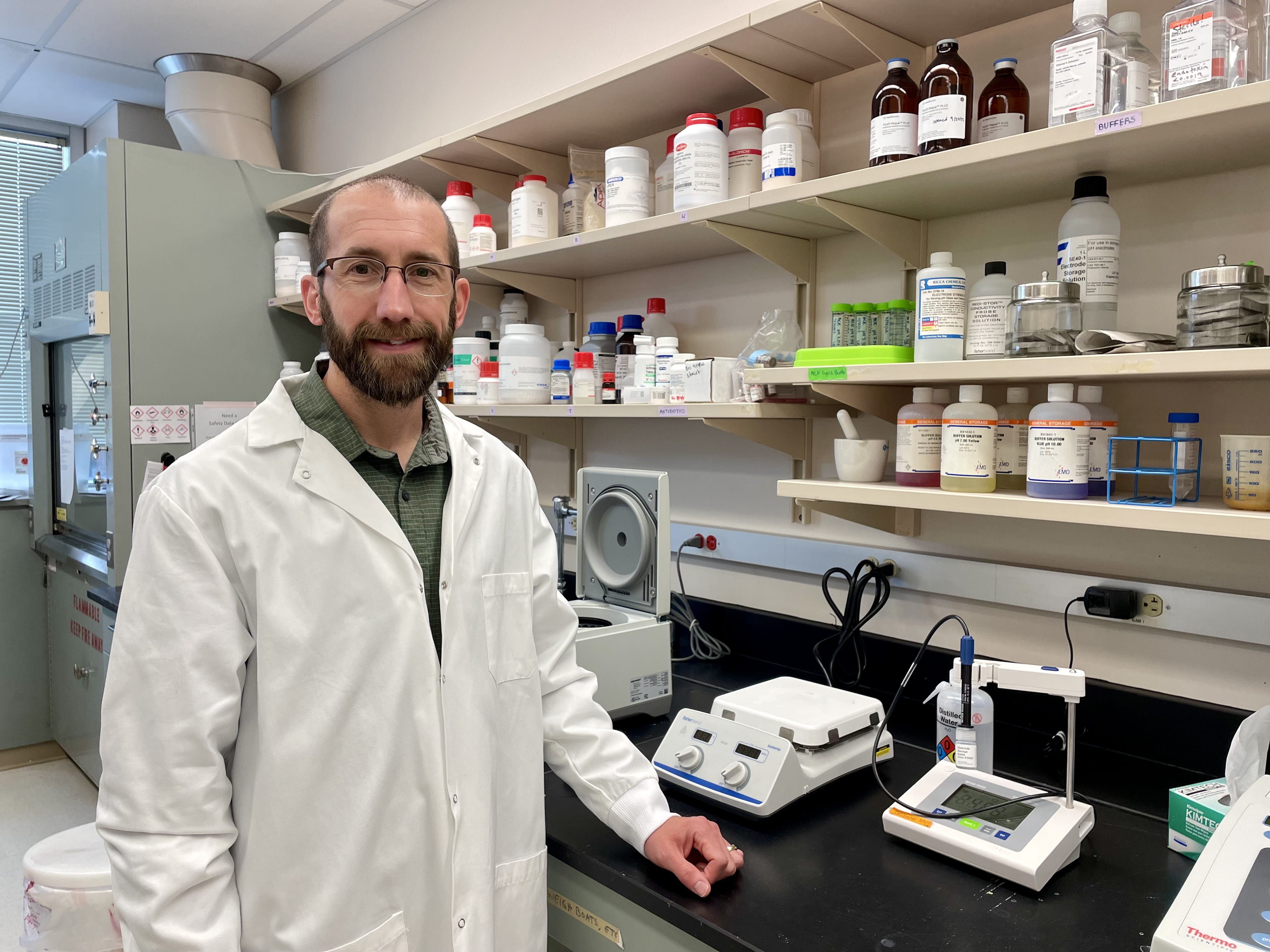 Nathan Schmidt wears a white lab coat and stands next to instrumentation in his lab