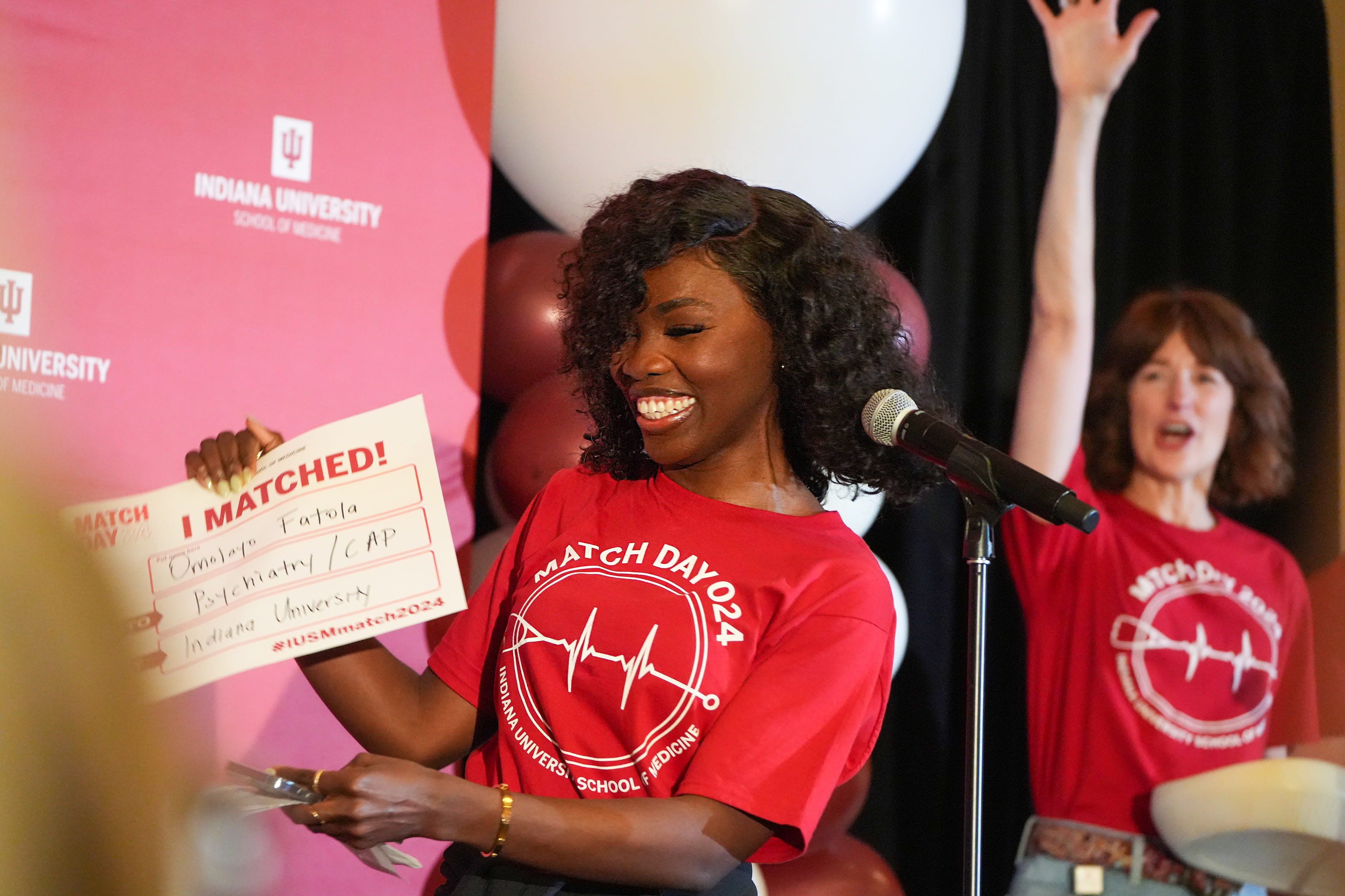 a student smiles and holds up her match day sign on stage. emily walvoord celebrates with her arms in the air in the background.