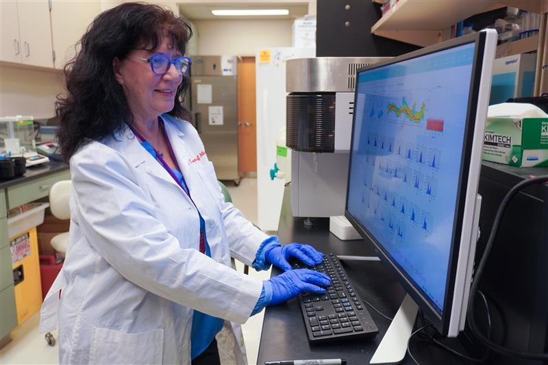 Joan Cook-Mills works at a computer station in her lab. She has long dark hair and is wearing glasses, a white lab coat and gloves.