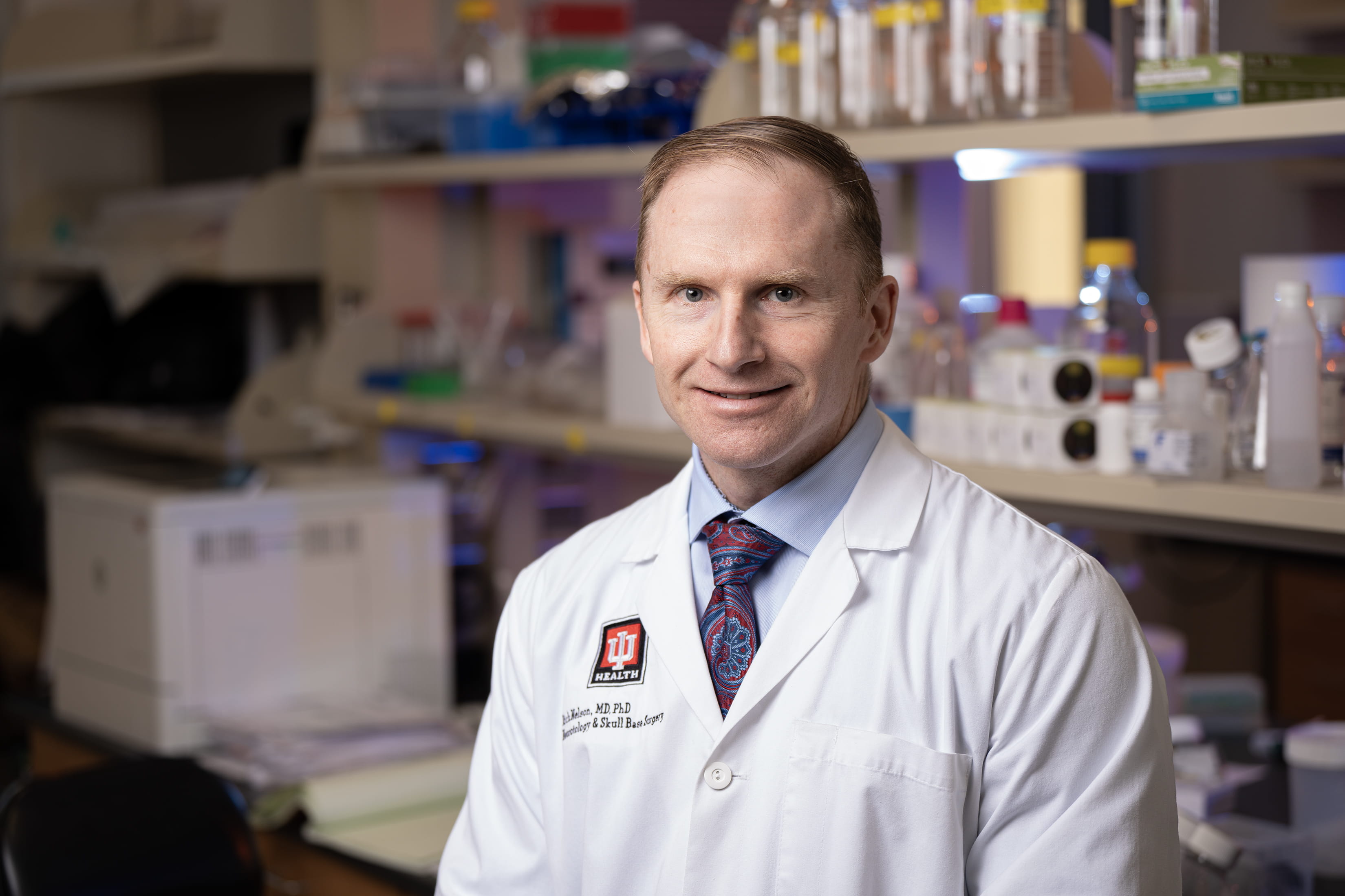 A man in a white lab coat poses in his lab. 