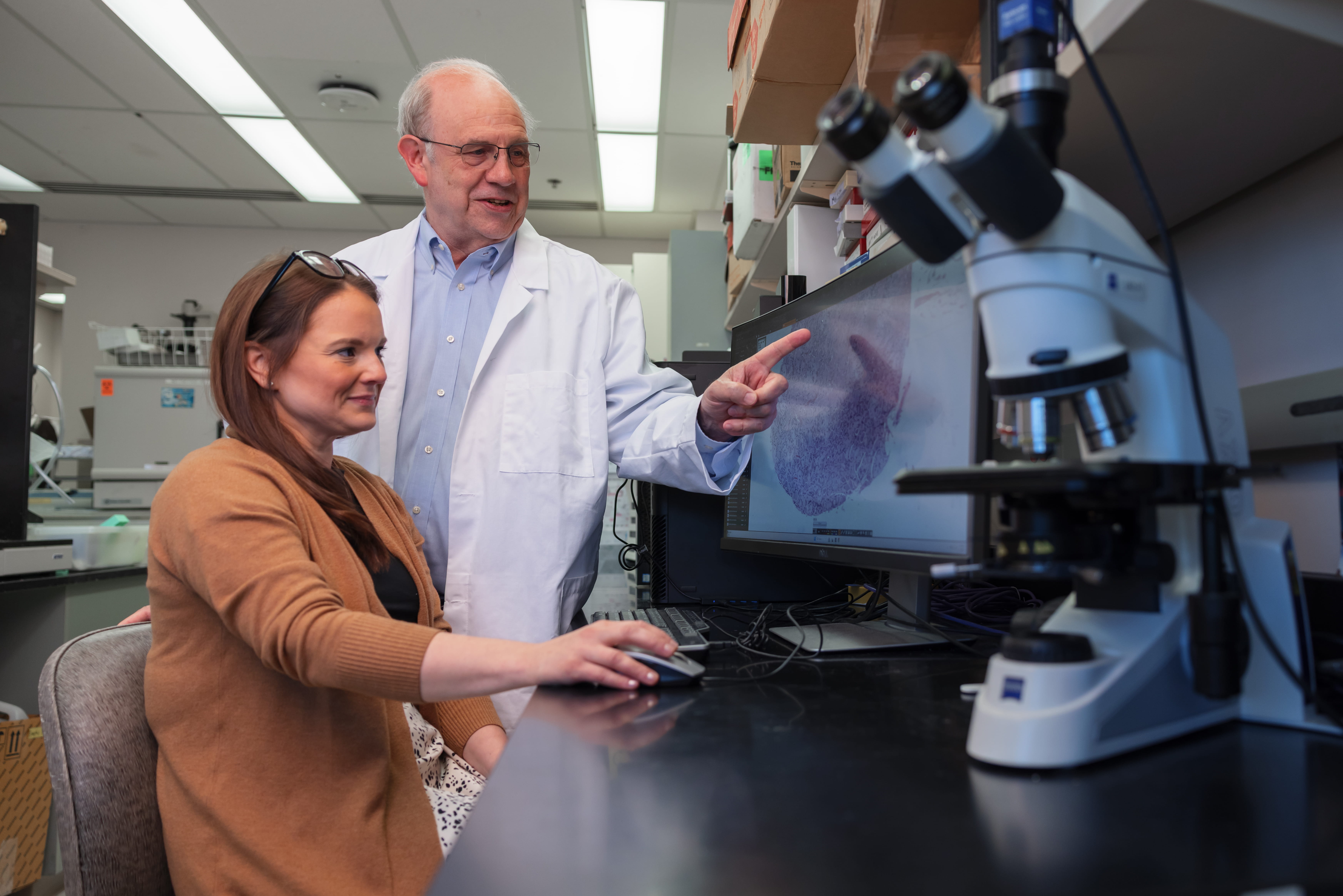 A man in a white lab coat watches as a woman uses a scientific tool.