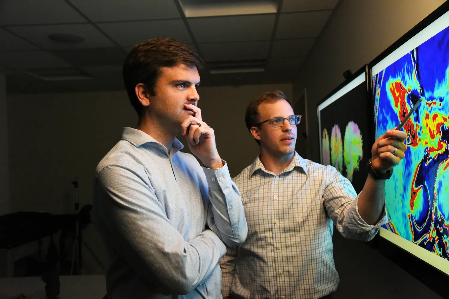Two men discussing colorful visual data on screens; one gestures while the other looks thoughtfully at the display.