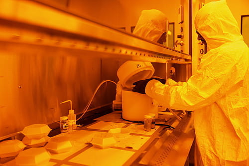A student in protective lab gear works under a hood in the lab, awash in orange light.
