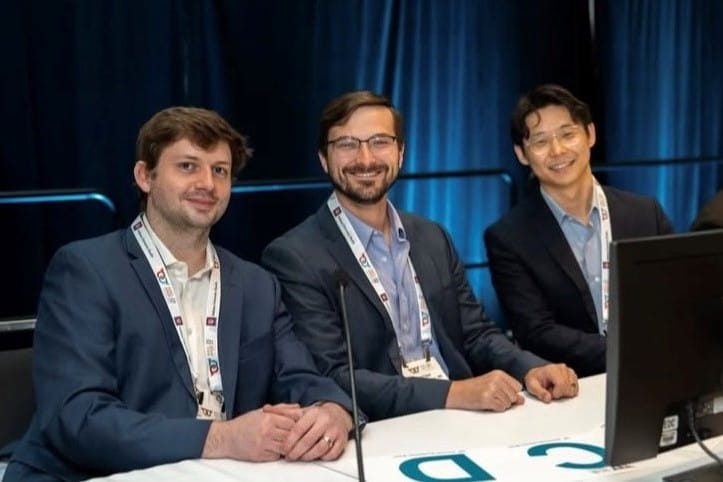Three individuals wearing conference badges sit at a table with microphones, facing the camera during a panel discussion.