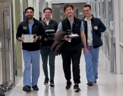 Four individuals walk down a hospital hallway carrying takeout food containers and trays. The group is dressed in a mix of scrubs, casual clothing, and jackets, with ID badges visible. The setting includes exam room doors along both sides of the corridor, an exit sign overhead.