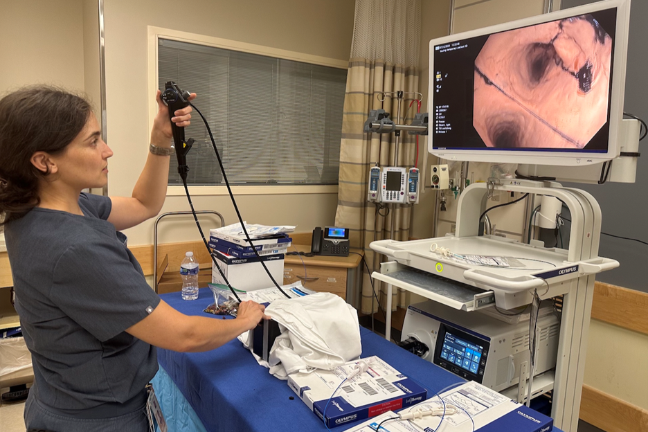 Healthcare professional preparing an endoscopy setup in a clinical room, holding an endoscope connected to a cart with medical equipment, while a monitor displays an internal view from the scope; procedure supplies and device packaging are arranged on a table nearby.