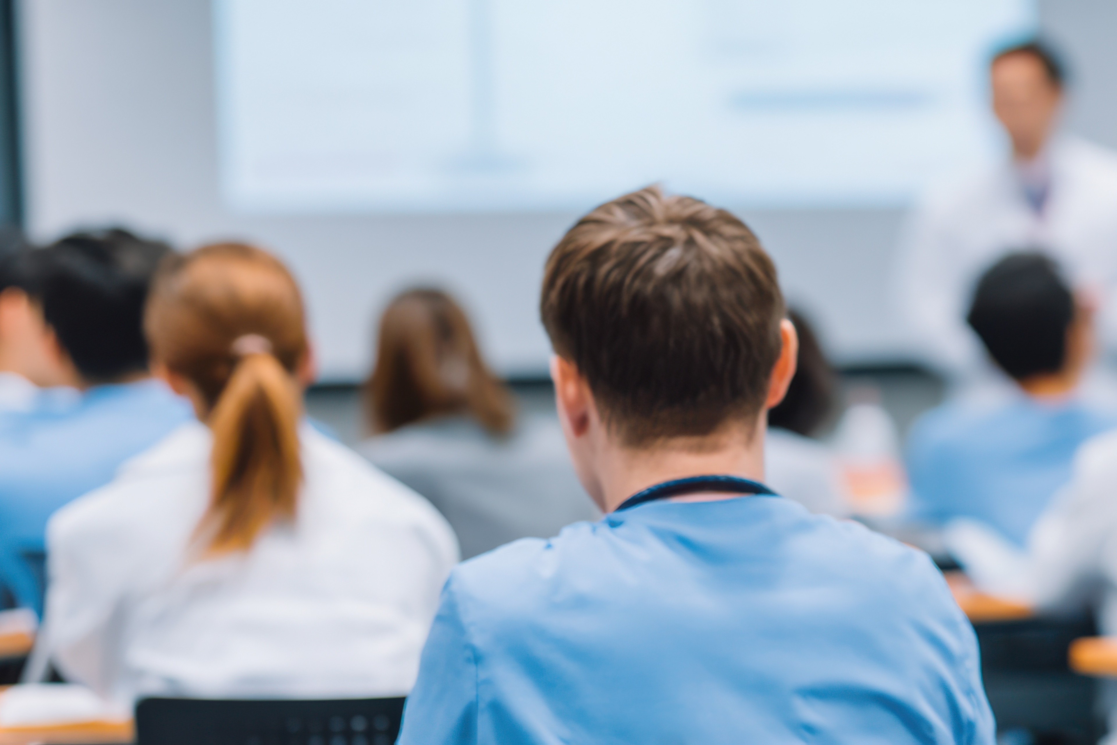 Group of people in a classroom facing forward while a presenter speaks at the front, with the foreground showing the back of a person in blue scrubs.