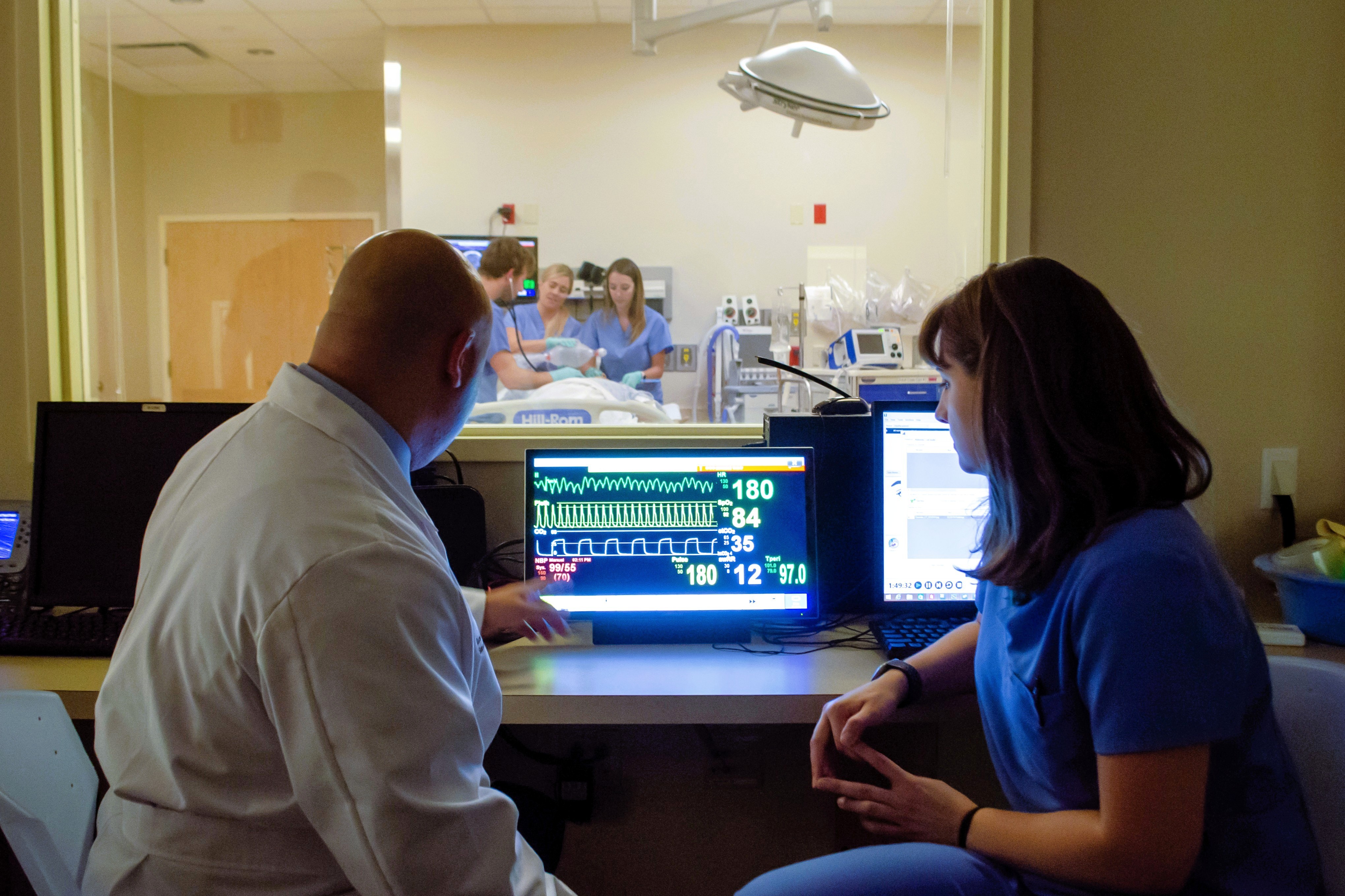 Clinicians review vital signs on a monitor while a team treats a patient in a simulation room behind glass.