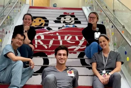 Five residents in scrubs sit on a hospital staircase with the Riley Hospital red wagon logo in the background