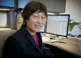 a researcher sits at her desk with a monitor filled with data in the background