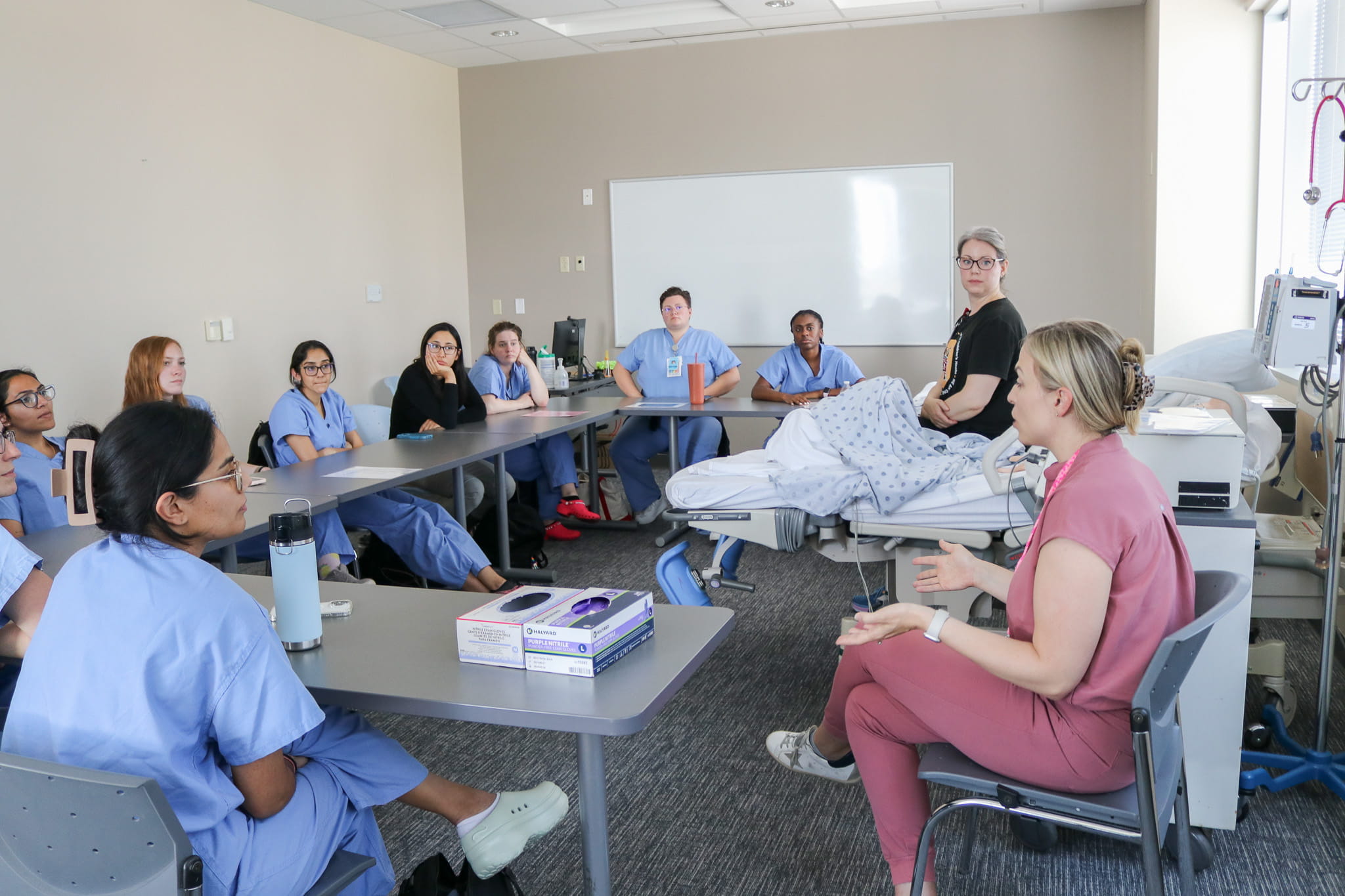 10 residents sit at a c-shaped table facing a patient and Emily Cassell, MD, at training