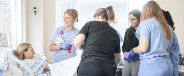 Three residents stand with two nurse educators around a simulated patient.