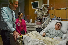 A male doctor speaks with a young patient in a hospital room. The patient is lying in a hospital bed. There is a woman in the background. 