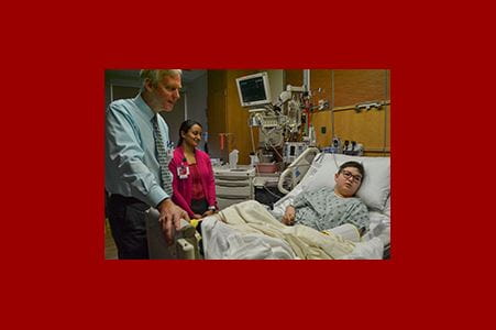 A male doctor speaks with a young patient in a hospital room. The patient is lying in a hospital bed. There is a woman in the background.