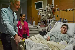 A male doctor speaks with a young patient in a hospital room. The patient is lying in a hospital bed. There is a woman in the background. 