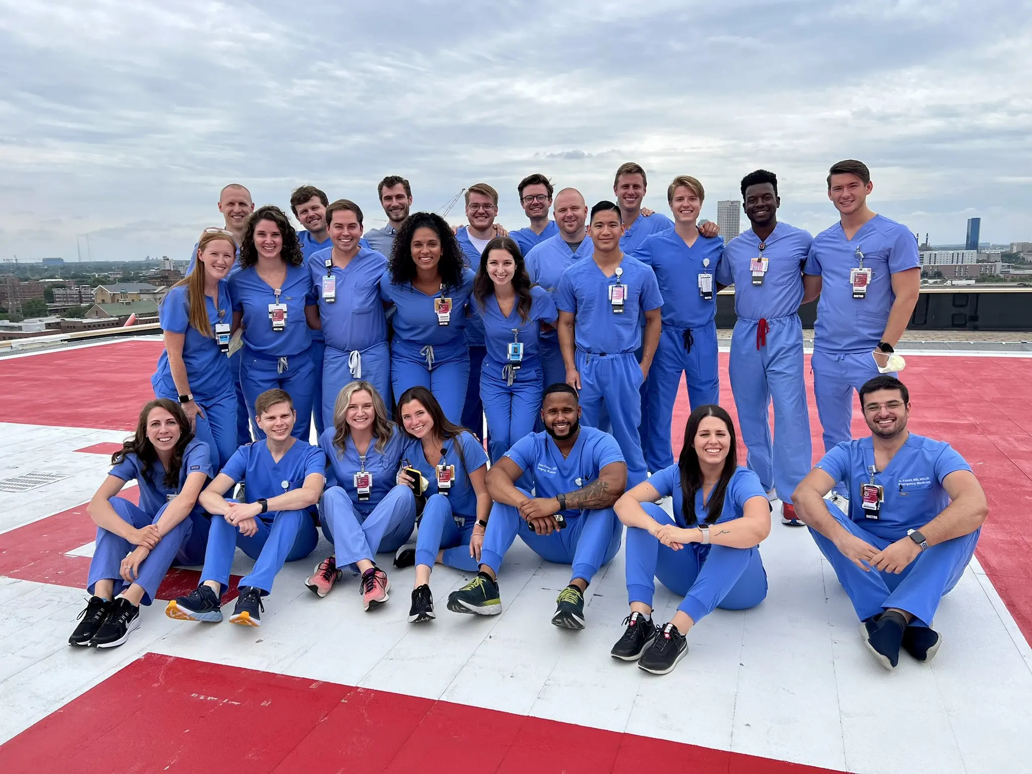 A group of emergency medicine residents gather on a red-and-white helipad for a group photo wearing their blue scrubs and IU Health badges