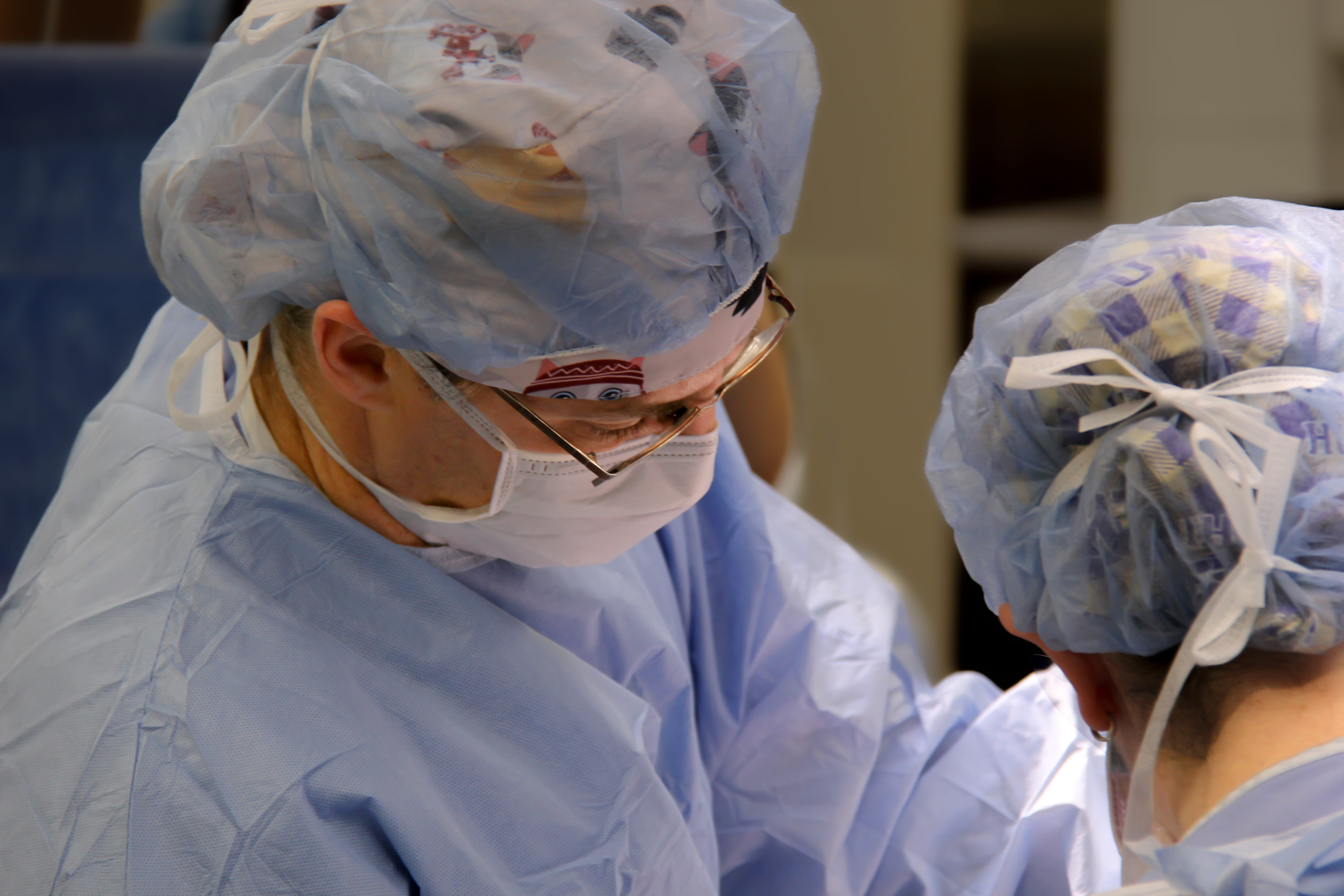 Roman Natoli stands facing an unseen surgery patient. He wears a gown, cap and surgical mask. The back of another surgery worker's head is visible.